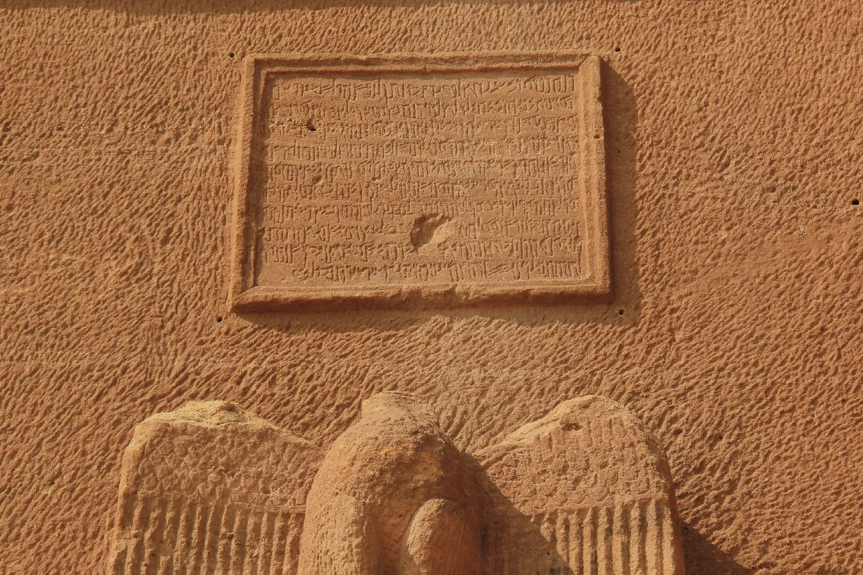 A sandstone facade with a framed panel of Nabataean inscription carved above two eagles with outspread wings.