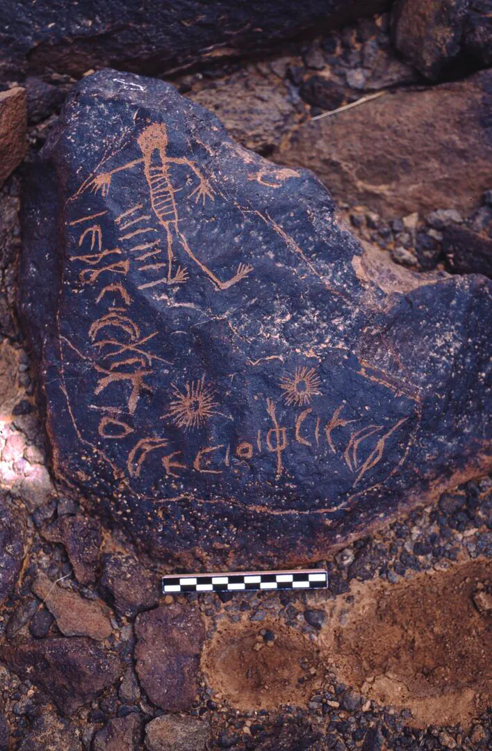 A dark basalt boulder incised with a Safaitic inscription, a stick figure and star motifs, with a photographic scale bar beside it.