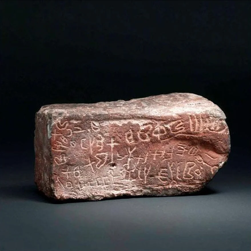 A rectangular red sandstone block inscribed with Thamudic letters, photographed against a dark background.
