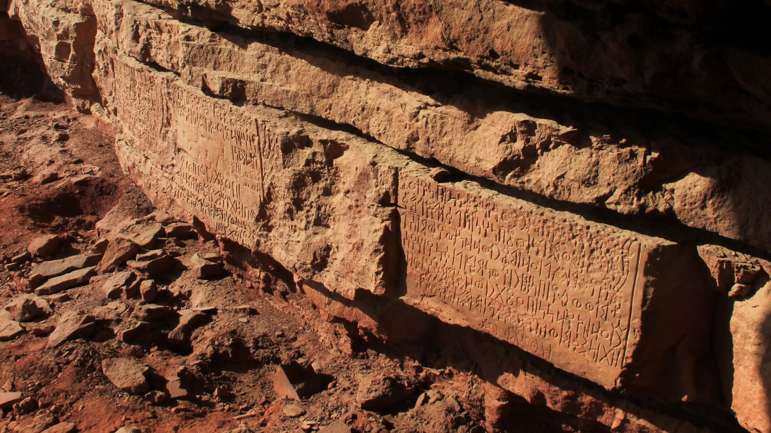 Sunlit sandstone ledges carved with long panels of Sabaic inscription, with rubble at their foot.