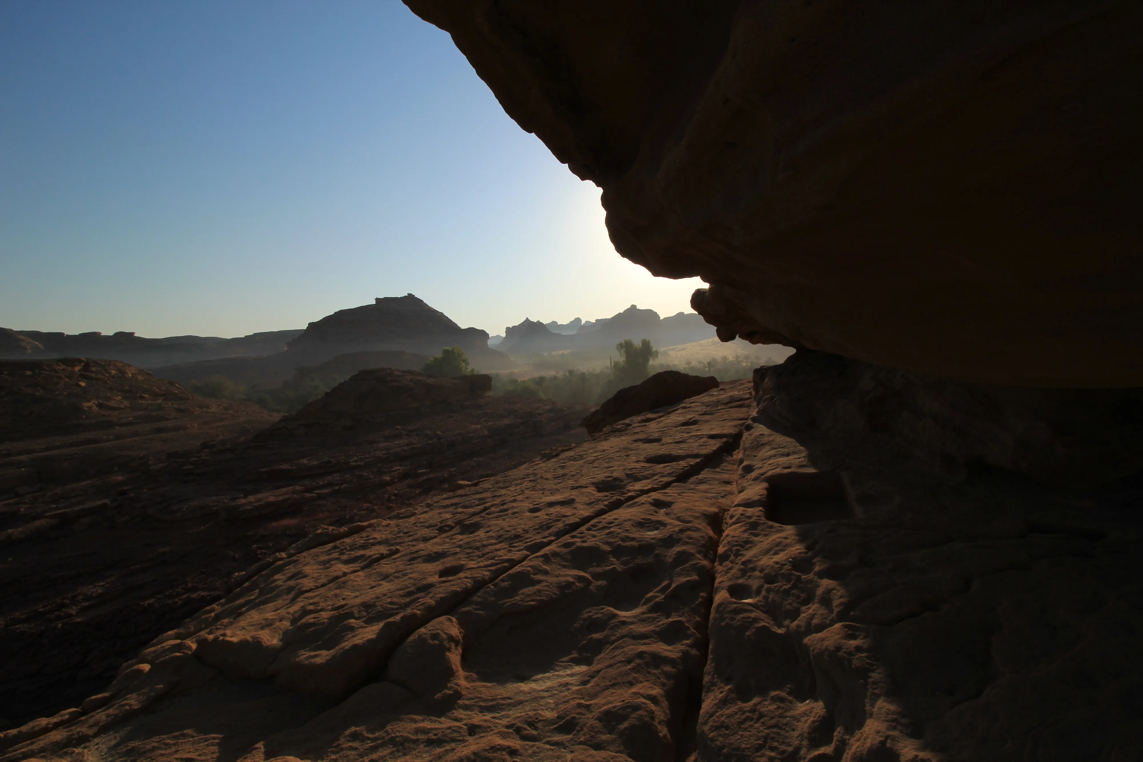 A view out from under a sandstone overhang, with hazy desert hills and palm groves catching the low sun.